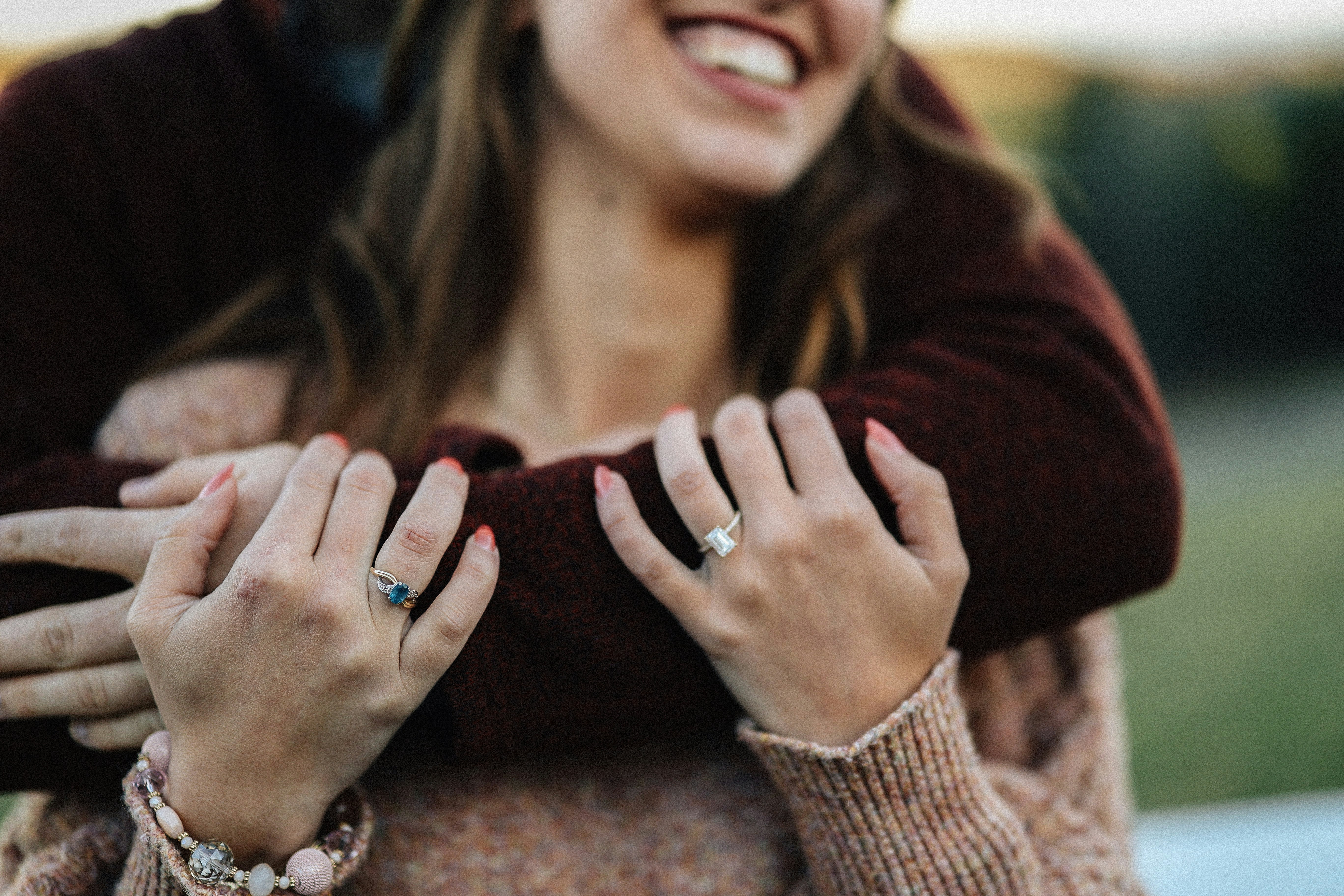 newly engaged couple, woman rings on both hands. fiance one arm around her hugging her from behind. Her arms grip his arm to her chest
