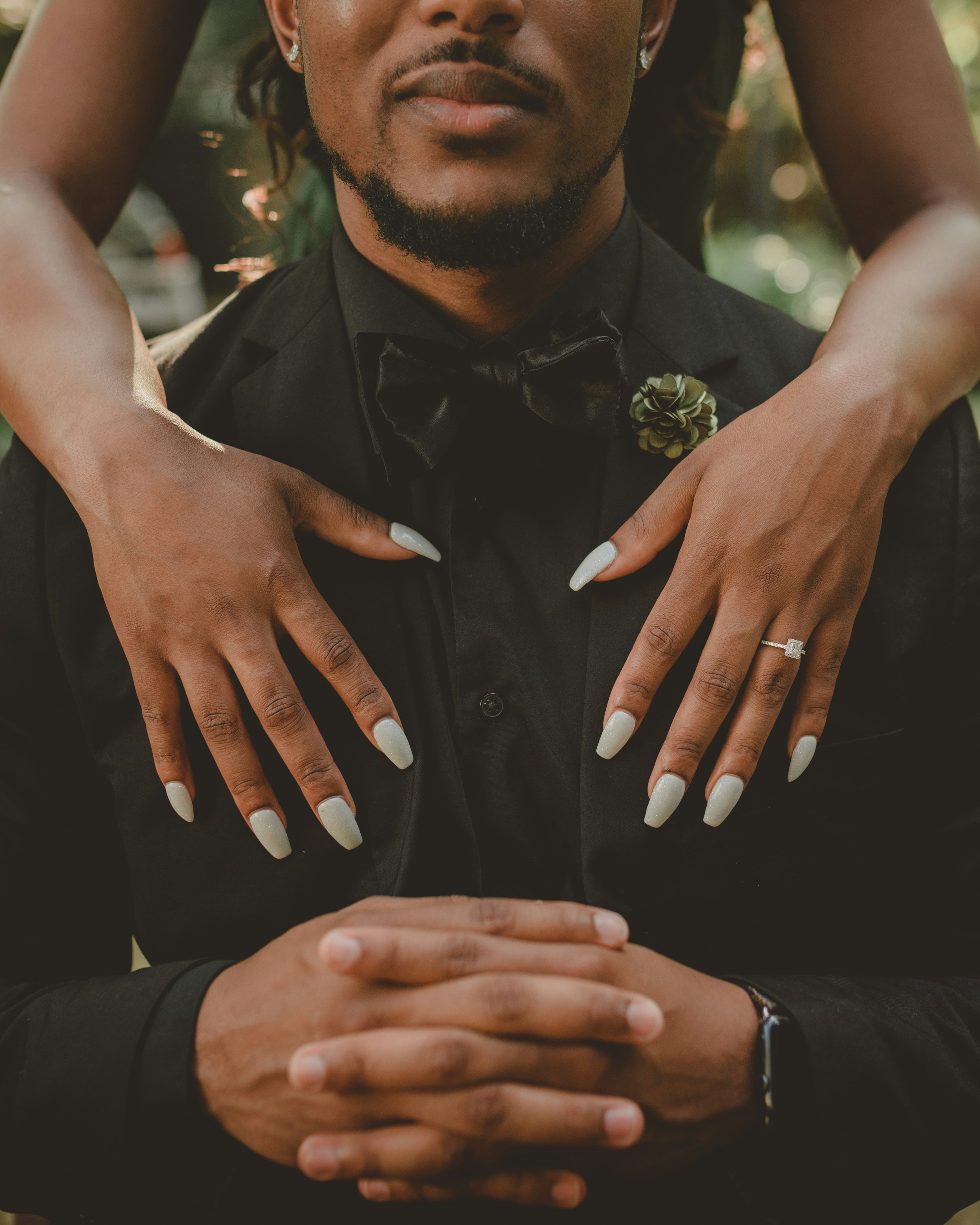powerful. man sitting, hands together wearing a black tux. Wife's hands Open standing behind him. Both hands on his chest   
